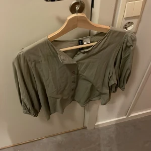 Olivgrön blus från Divided - Säljer en olivgrön blus från Divided med puffiga långärmar och knappar framtill. Blusen har en croppad passform och är tillverkad i mjukt, tunt material som ger en skön känsla. Perfekt för dig som gillar stilrena och trendiga plagg.