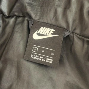 Svart pufferjacka från Nike med huva - Svart pufferjacka från Nike med vit logga på bröstet och coola swoosh-detaljer på huvan och nederkanten. Jackan har dragkedja framtill, snörning i huvan och är quiltad för extra värme. Perfekt för dig som gillar sportig streetstyle.