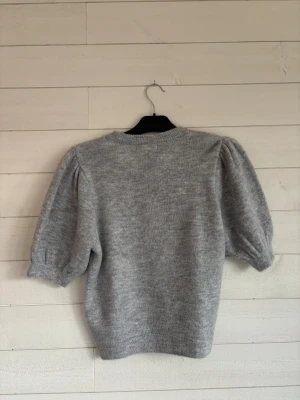 Grå stickad tröja med puffärm - Mysig grå stickad tröja med puffiga korta ärmar och rund hals. Tröjan har ribbade muddar vid hals, ärmslut och nederkant. Perfekt för dig som gillar enkel men trendig stil och vill ha något som känns både chill och snyggt.