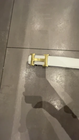 Vit skinnbälte med guld H-spänne - Snyggt vitt bälte i skinn med ett stort, stilrent H-format spänne i guld. Bältet har en klassisk rak form och är perfekt för att lyfta din outfit med en exklusiv touch. Passar dig som gillar statement-accessoarer och vill sticka ut med något lyxigt.