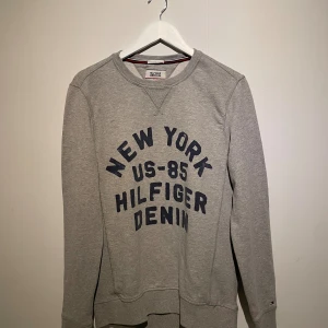 Grå Hilfiger Denim sweatshirt M - Grå sweatshirt från Hilfiger Denim med mörkblå tryck på bröstet där det står 'New York US-85 Hilfiger Denim'. Tröjan har rund hals, långa ärmar och ribbade muddar. Perfekt för dig som gillar enkel och sportig stil.