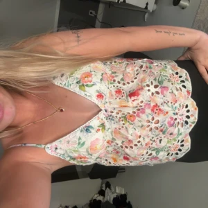 Blommig vit linnetopp med spetsdetaljer - Superfin linnetopp med blommigt mönster i rosa, rött, grönt och orange på vit botten. Toppen har tunna axelband, v-ringning och spetsdetaljer med broderade hålmönster. 