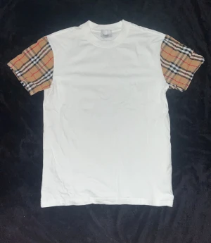  Burberry t-shirt   - Snygg vit t-shirt från Burberry med klassiska rutiga ärmar i beige, svart, vitt och rött. T-shirten har rund hals och är tillverkad i mjuk bomull. Perfekt för dig som vill ha en stilren look med en touch av lyx.