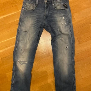 Replay Anbass blå jeans med slitningar - Snygga Replay Anbass jeans i blå tvätt med slitna detaljer och lätt blekta partier. Klassisk femficksmodell med raka ben och normal passform. Jeansens material är bomull och de har Replay-logga på bakfickan samt läderpatch i midjan.