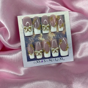 FrenchGold(S):Medium Square - Snygga handgjorda press on-naglar i fransk stil med nude bas och handmålade vita tippar, dekorerade med guldiga pärlor och söta rosetter med pärldetalj. Naglarna är medium längd och har en glansig finish, perfekta för dig som vill ha en extra glam look.S: 15 mm, 11,5 mm, 12 mm, 11 mm, 9 mm  