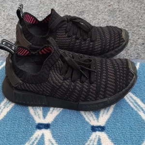 Svarta Adidas NMD sneakers - Säljer ett par svarta Adidas NMD sneakers med stickad ovandel och diskret mönster. Skorna har svarta snören, svart sula och detaljer i plast vid hälen. Insidan har rosa ränder för en extra touch. Perfekta för dig som gillar streetstyle och bekväma skor.