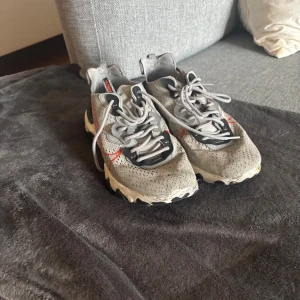 Nike React Element 55 grå sneakers - Nike React Element 55 sneakers i grått med svarta och vita detaljer samt en röd swoosh på sidan. Ovandelen är i syntetmaterial med perforerat mönster och snörning framtill. Sulan är vit med svarta partier och har en vågig struktur för extra grepp.