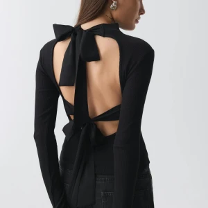 Svart ribbad långärmad topp med rosett - Svart långärmad topp med öppen rygg. Toppen har två stora rosetter i ryggen, en vid nacken och en vid midjan, som ger en unik look. Materialet är mjukt och stretchigt, perfekt för att sticka ut. Snygg att matcha med svarta jeans eller kjol. Är i storlek M men funkar jätte bra om man har S