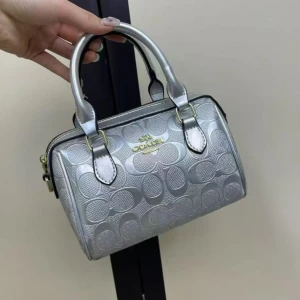 Coach handväska i olika färger - Snygg handväska från Coach med präglat logomönster och guldfärgade detaljer. Finns i silver, vinröd, svart och vit. Väskan har två handtag, dragkedja och avtagbar axelrem. Materialet är glansig syntet med exklusiv känsla. Perfekt storlek för det viktigaste.