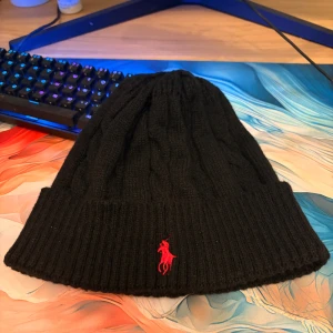 Polo Ralph Lauren mössa - Svart kabelstickad mössa från Polo Ralph Lauren med röd broderad logga framtill. Tillverkad i 100% bomull och har bred ribbad kant. Perfekt för dig som vill ha en klassisk och stilren look med en touch av street.