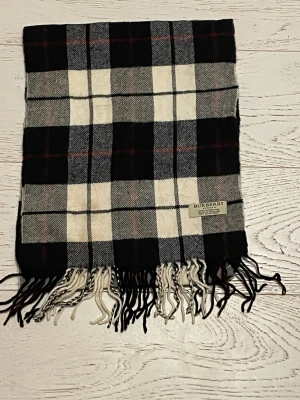 Rutig halsduk från Burberry i ull - Snygg halsduk från Burberry med klassiskt rutmönster i svart, vit och grå. Gjord av 100% lammull och har fransar i ändarna. Perfekt för att ge din outfit en lyxig touch och hålla dig varm under kyliga dagar.