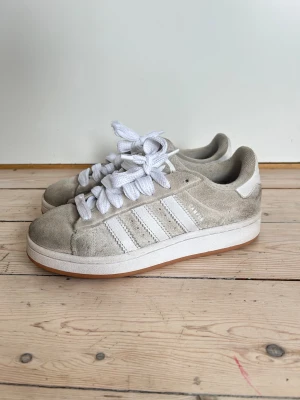 Beige Adidas Campus sneakers mocka - Säljer ett par  Adidas Campus sneakers i beige mocka med klassiska vita ränder och grova vita snören. Skorna har en platt sula i gummi och rund tå, perfekt för dig som gillar retrovibe och enkel design. Campus-loggan syns på sidan.