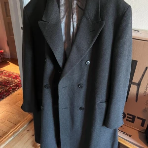 Mörkgrå rock från Fellini i ull/kashmir - Snygg mörkgrå rock från Fellini med dubbelknäppning och klassisk krage. Tillverkad i en lyxig blandning av ull och kashmir, fodrad med grått tyg. Perfekt för dig som vill ha en stilren och tidlös look under höst och vinter. Passar L/XL