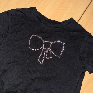 Svart T-shirt  med rosett XS - Svart kortärmad tshirt med rund hals och stor rosett framtill, dekorerad med glittrande strass-stenar. Tröjan i grund är från lager 157, går att tvätta OBS! Bara i 30 grader, ingen torktumling 