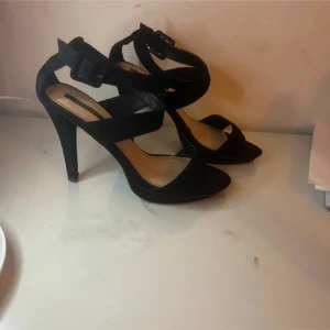 Svarta klackar med remmar - Snygga svarta sandaler med hög klack och breda remmar i mockaimitation. Skorna har öppen tå, justerbart spänne vid ankeln och en elegant, spetsig siluett. Perfekta för dig som vill ha en stilren och trendig look. 9cm klack.