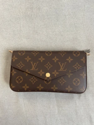 Louis Vuitton Monogram axelväska - Snygg brun axelväska från Louis Vuitton med klassiskt monogrammönster och guldfärgad kedja. Väskan har ett kuvertformat lock med knappstängning och kommer med matchande plånbok och pouch. Perfekt för dig som vill ha en ikonisk designeraccessoar.