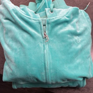 Turkos velourjacka från Juicy Couture - Säljer en turkos velourjacka från Juicy Couture med dragkedja och snörning i huvan. Jackan har ett broderat JC-logo på bröstet och ribbade muddar. Perfekt för chill dagar och streetstyle.