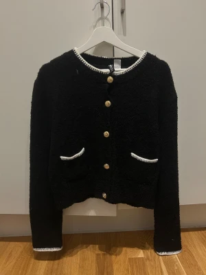 Svart kofta med vita detaljer från H&M - Snygg svart kofta från H&M med vita broderade detaljer vid krage, fickor och ärmslut. Guldiga knappar framtill och två små fickor. Mjuk och fluffig känsla, perfekt för att lyfta din outfit.