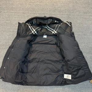 Svart jacka från Burberry - Finns storlek S  pris kan diskuteras Snygg svart dunjacka från Burberry med klassiskt rutigt foder i huvan. Jackan har hög krage, dragkedja och knappar framtill samt två dragkedjeförsedda fickor. Perfekt för kalla dagar och har en riktigt lyxig känsla.