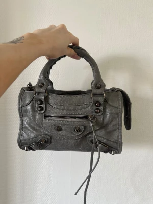 Grå handväska  - Snygg grå handväska i mjukt skinn med coola nitar och detaljer. Väskan har dubbla handtag, dragkedja framtill och långa dekorativa remmar. Axelband finns till. Köpt på arkivet för 800 kr