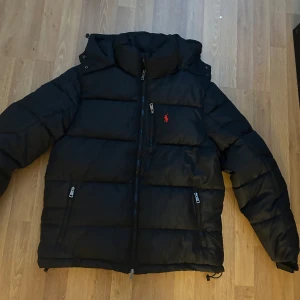 Svart pufferjacka från Polo Ralph Lauren - Svart pufferjacka från Polo Ralph Lauren med röd logga på bröstet. Jackan har huva, flera dragkedjefickor och quiltad design. Materialet är polyester och bomull, vilket gör den både lätt och varm. Perfekt för kalla dagar och har en snygg, modern look.