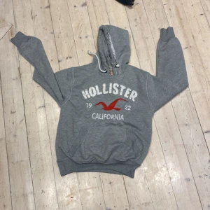 Grå hoodie från Hollister - Säljer en grå hoodie från Hollister med stor vit logga och röd fågel på bröstet. Tröjan har huva med snörning och en stor magficka. Perfekt för chill dagar och har en klassisk streetstil. Passar dig som gillar avslappnat och snyggt.
