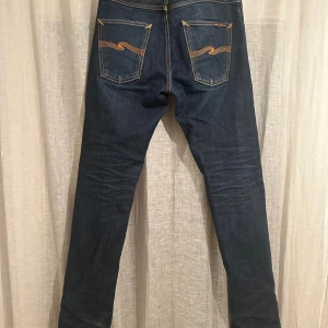 Mörkblå jeans från Nudie Jeans Co - Snygga mörkblå jeans från Nudie Jeans Co med klassisk orange kontrastsöm och broderad våg på bakfickorna. Modellen har fem fickor, knapp och dragkedja fram samt läderpatch med logga bak. Jeansen har en rak och smal passform som sitter riktigt bra.