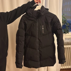 Svart pufferjacka från Sail Racing - Svart pufferjacka från Sail Racing med huva och flera praktiska dragkedjefickor framtill. Jackan har en stilren look med logga på bröstet och ett märke på ärmen. Långärmad och med en skön, lite längre passform som håller dig varm under kalla dagar.