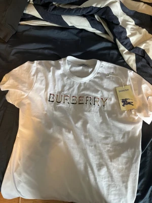 Vit Burberry t-shirt med broderad logga - Snygg vit t-shirt från Burberry i mjuk bomull. Framtill finns en broderad Burberry-logga med klassiskt rutmönster i bokstäverna och texten 'London England' under. T-shirten har rund hals och korta ärmar, perfekt för dig som gillar stilrena designer med lyxig touch.