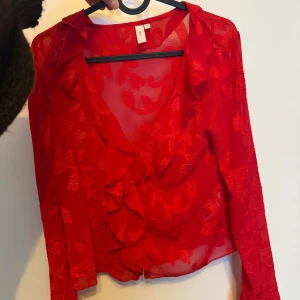 Röd blus med volang och broderat mönster - Säljer en transparent röd blus från Nelly med broderat blommönster och volang längs v-ringningen. Blusen har långa ärmar och är lätt croppad framtill. Använd max 2 gånger 