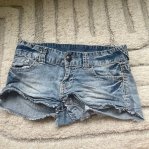 Blå jeansshorts från SisterS Point - Snygga blå jeansshorts från SisterS Point med låg midja och fransig kant. Klassisk femficksmodell med slitna detaljer och tydliga kontrastsömmar. Perfekt för varma dagar och chill stil. Knapp och dragkedja framtill.