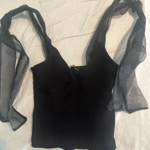 Svart ribbad topp med meshband från Zara - Säljer en svart ribbad topp från Zara med bred v-ringning och långa meshband som kan knytas på axlarna. Toppen är croppad och har en tight passform. Materialet är mjukt och stretchigt, och meshdetaljerna ger en cool kontrast till den ribbade ytan.
