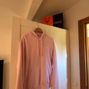 Ljusrosa hoodie från Nike - Säljer en ljusrosa hoodie från Nike med broderad vit logga på bröstet. Tröjan har huva med snörning, känguruficka och långa ärmar. Perfekt för dig som gillar en chill och sportig stil.