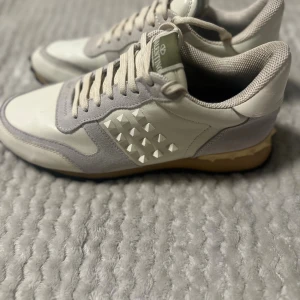 Valentino Rockrunner sneakers beige/vit - Snygga Valentino Rockrunner sneakers i beige och vitt med coola nitar på sidorna och hälen. Skorna har en mix av skinn och mocka, snörning och en platt sula. Perfekta för dig som gillar exklusiva och trendiga detaljer.