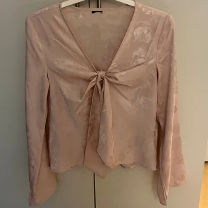 Ljusrosa blus med knyt och blommönster - Supersöt ljusrosa blus med subtilt blommönster och lång ärm. Toppen har v-ringning och snygg knytning framtill som ger en feminin touch. Materialet är glansigt och känns lyxigt, perfekt för att lyfta vilken outfit som helst.