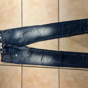 Replay Anbass jeans blå slim fit - Snygga Replay Anbass jeans i blå tvätt med slitningar och detaljerad faded effekt. Slim fit-modell med klassiska fem fickor, dragkedja och knappstängning. Märkespatch bak i brunt och Replay-logga vid fickan. Perfekt passform för en modern look.