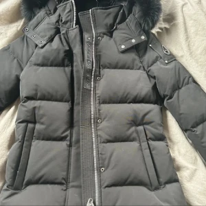 Svart dunjacka Moose Knuckles M - Säljer en svart dunjacka från Moose Knuckles i storlek M. Jackan har puffer-design, avtagbar huva med fuskpäls, dragkedja och snygga detaljer med logga på ärmen. Perfekt för kalla vinterdagar och riktigt stilren look.