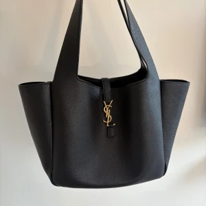 YSL Saint Laurent Le 5 à 7 Bea - Stilren svart shoppingväska från Saint Laurent i snyggt skinn med guldigt YSL-emblem framtill. Väskan har breda handtag, rymligt fack och en innerficka med dragkedja, går även att ”knyta” så den blir mindre. Äkta, köptes från vestiarie collective, serienummer se sista bild. Loggan har tappat lite färg, men annars jättefint skick🖤ENDAST SERIÖSA KÖPARE