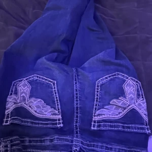 Blå jeans med broderade bakfickor💙 - Snygga blå jeans med kontrastsömmar och unika broderade detaljer på bakfickorna. Klassisk femficksmodell med knapp och dragkedja framtill. Jeansen har en utsvängd passform från ginatrickot 🩷