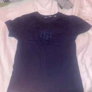 Mörkblå GANT t-shirt med broderad logga - Snygg mörkblå t-shirt från GANT med broderad logga och sköld på bröstet. Klassisk rund hals och korta ärmar. Tillverkad i mjuk bomull som känns skön mot huden. Perfekt för en clean och sportig look.