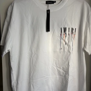 Vit AMIRI t-shirt med färgstänkt tryck - Snygg vit t-shirt från AMIRI i bomull med coolt tryck framtill där loggan har färgstänk och droppande effekt i svart, orange, blått och mint. Klassisk passform och rund hals. Perfekt statement-plagg för dig som gillar streetstyle.