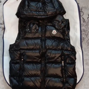 Svart dunväst från Moncler med huva - Säljer en svart, glansig dunväst från Moncler med avtagbar huva och två dragkedjeförsedda fickor. Västen har ett quiltat mönster och Moncler-logga på bröstet. Perfekt för lager-på-lager och snygg streetstil. Nfc funkar