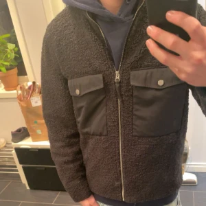 Svart teddyjacka från These Glory Days - Mysig svart teddyjacka från These Glory Days med två stora bröstfickor i nylon och dragkedja framtill. Jackan har hög krage och en schysst, lite boxig passform. Perfekt för kalla dagar när du vill vara både varm och snygg. Skick 9/10