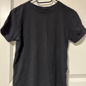 Svart t-shirt från Kappahl - En basic svart t-shirt från Kappahl i storlek 158/164. T-shirten har rund halsringning och korta ärmar, tillverkad i mjuk bomull. Perfekt till jeans eller shorts för en clean och enkel look.