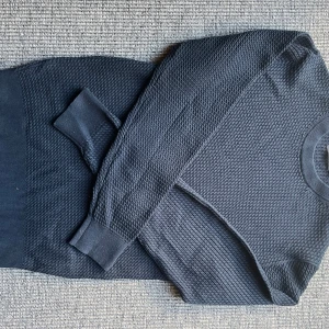 Mörkblå stickad tröja Dressmann S - Mörkblå stickad tröja från Dressmann i storlek S. Tröjan har ett diskret strukturerat mönster, rund halsringning och ribbade muddar vid ärmslut och nederkant. Perfekt för lager-på-lager under kyliga dagar.