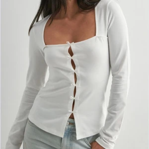 Vit långärmad topp med snörning - Säljer en vit långärmad topp med fyrkantig urringning och snygga rosetter framtill. Toppen har en figurnära passform. Perfekt att styla med jeans eller kjol för en trendig look.