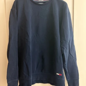 Mörkblå sweatshirt från Tommy Jeans - Mörkblå sweatshirt från Tommy Jeans i relaxed fit. Tröjan har rund halsringning, långa ärmar och en broderad logga nertill. Tillverkad i mjuk bomull som är skön att ha på sig. Perfekt för dig som gillar en enkel och stilren look.