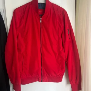 Röd bomberjacka från Selected Homme - Snygg röd bomberjacka från Selected Homme med klassisk ribbad krage och muddar. Jackan har dragkedja framtill och en extra ficka med zip på ärmen. Perfekt för dig som vill sticka ut med färg och stil.