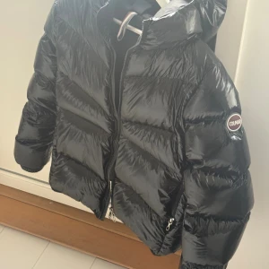 Svart pufferjacka från Colmar - Säljer en svart, glansig pufferjacka från Colmar Originals med huva och dragkedja framtill. Jackan har ett stilrent, quiltat mönster och logotyp på ärmen. Perfekt för kalla dagar. Säljer då den ej kommer till användning. Den är i nyskick och det finns inga tecken på användning. Nypris -5000