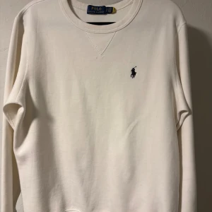 Vit sweatshirt från Polo Ralph Lauren - Vit sweatshirt från Polo Ralph Lauren med klassisk rund hals och broderad marinblå logga på bröstet. Tröjan har långa ärmar och är tillverkad i mjuk bomull, perfekt för en clean och stilren look.